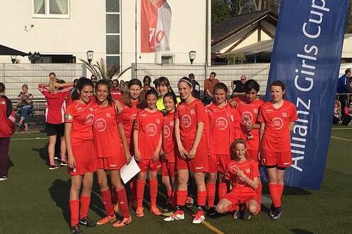 Gruppe junger Fu&szlig;ballerinnen in roten Trikots posiert auf einem Sportplatz mit Allianz Girls Cup-Banner im Hintergrund.