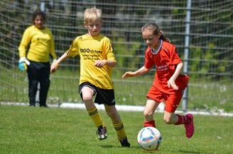 Zwei Kinder spielen Fu&szlig;ball auf einem Platz, ein Junge in gelbem Trikot und ein M&auml;dchen in rotem Trikot.