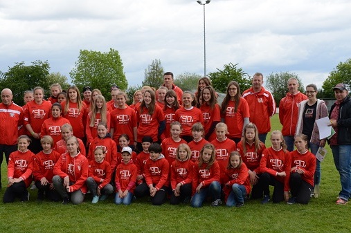 Gruppensfoto von 40 Personen in roten Shirts auf einem Sportplatz, umgeben von gr&uuml;nen B&auml;umen unter bew&ouml;lktem Himmel.