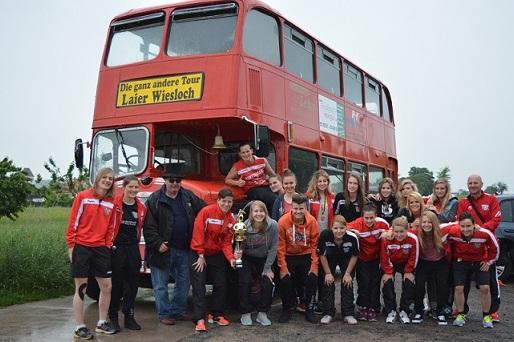 Gruppe von Personen in Sportkleidung posiert vor einem roten Doppeldeckerbus, einige halten einen Pokal.