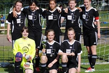 Mädchenfußballmannschaft in schwarzen Trikots mit Medaillen und Pokal auf einem Fußballplatz, umgeben von einem Tor.