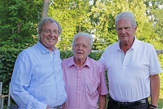 Drei M&auml;nner stehen l&auml;chelnd im Freien vor gr&uuml;ner Hintergrundvegetation, tragen legere Hemden und posieren nah beieinander.