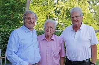 Drei M&auml;nner stehen l&auml;chelnd im Freien vor gr&uuml;ner Hintergrundvegetation, tragen legere Hemden und posieren nah beieinander.