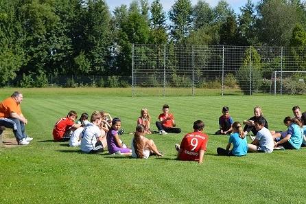 Gruppe von Kindern und Jugendlichen sitzt im Kreis auf einer Wiese, w&auml;hrend ein Erwachsener auf einer Bank zuschaut.