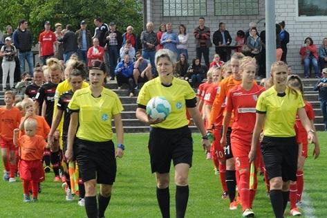 Schiedsrichterinnen und Spielerinnen betreten das Spielfeld, umgeben von Zuschauern auf einer Sportveranstaltung.