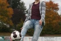 Eine Frau jongliert mit einem Fu&szlig;ball auf einem Rasenplatz, umgeben von herbstlich gef&auml;rbten B&auml;umen.