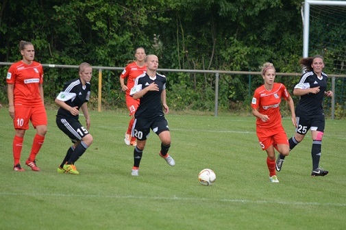F&uuml;nf Fu&szlig;ballspielerinnen, zwei in Rot und drei in Schwarz, laufen auf einem Rasenfeld einem Ball entgegen.
