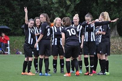 Eine Gruppe von acht Fu&szlig;ballspielerinnen in schwarz-wei&szlig;en Trikots posiert jubelnd auf einem Rasenplatz.