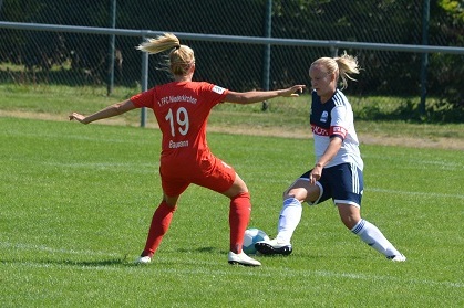 Zwei Fu&szlig;ballspielerinnen im Wettkampf um den Ball auf einem Rasenplatz, eine in rotem, die andere in wei&szlig;-blauem Trikot.