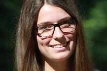 Portr&auml;t eines l&auml;chelnden M&auml;dchens mit langen Haaren, Brille und Perlenkette, vor einem unscharfen, gr&uuml;nen Hintergrund.