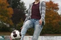 Eine Frau jongliert mit einem Fu&szlig;ball auf einem Rasenplatz, umgeben von herbstlich gef&auml;rbten B&auml;umen.