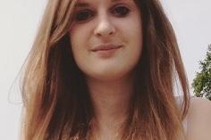 Junge Frau mit langen, braunen Haaren und l&auml;chelndem Gesicht, stehend vor einem unscharfen Hintergrund.