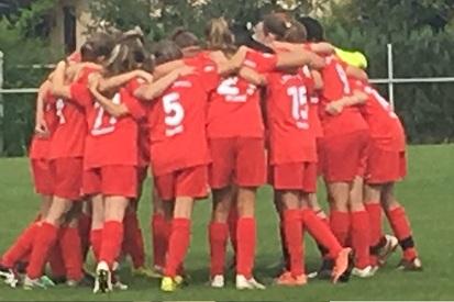 Mannschaft von Frauen in roten Trikots umarmt sich im Kreis auf einem Fu&szlig;ballplatz vor einem Spiel.