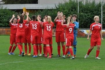 Frauenfußballmannschaft in roten Trikots feiert ein Tor auf dem Spielfeld, umgeben von grüner Wiese und Zuschauerbereich.