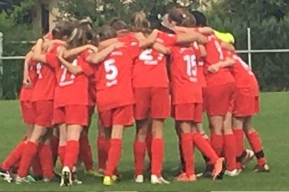 Mannschaft von Frauen in roten Trikots umarmt sich im Kreis auf einem Fu&szlig;ballplatz vor einem Spiel.