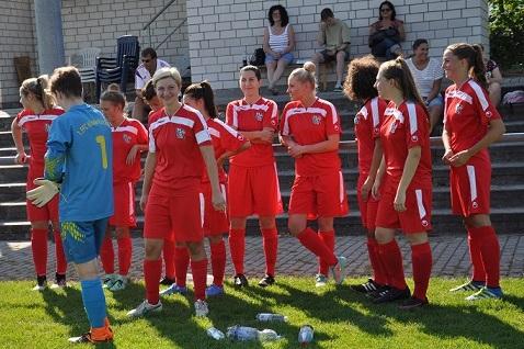 Ein Fu&szlig;ballteam in roten Trikots steht am Spielfeldrand, w&auml;hrend die Torh&uuml;terin ihren Knoten bindet. Zuschauer im Hintergrund.