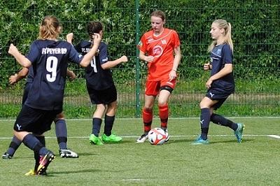 F&uuml;nf M&auml;dchenfu&szlig;ballspielerinnen auf einem Kunstrasenfeld, eine in rot dribbelt den Ball, vier in dunkelblau greifen an.