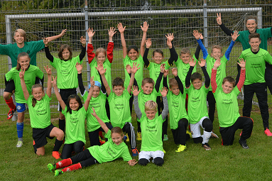 Gruppe von Kindern in gr&uuml;nen Trikots mit erhobenen H&auml;nden, umgeben von einem Fu&szlig;ballfeld und einem Tor.
