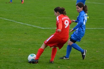 Zwei Fu&szlig;ballspielerinnen im Wettkampf, eine in rotem und die andere in blauem Trikot, auf einem gr&uuml;nen Spielfeld.