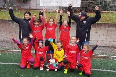 Mädchenfußballmannschaft in roten Trikots posiert fröhlich vor einem Tor, Trainer und Spielerinnen heben die Arme.