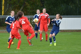 Zwei Frauenteams in roten und blauen Trikots spielen Fu&szlig;ball auf einem gr&uuml;nen Platz, ein Ball fliegt zwischen ihnen.