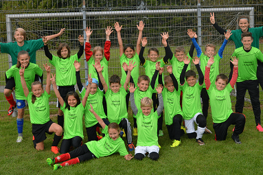Gruppe von Kindern in gr&uuml;nen Trikots mit erhobenen H&auml;nden, umgeben von einem Fu&szlig;ballfeld und einem Tor.