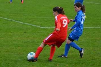 Zwei Fu&szlig;ballspielerinnen im Wettkampf, eine in rotem und die andere in blauem Trikot, auf einem gr&uuml;nen Spielfeld.