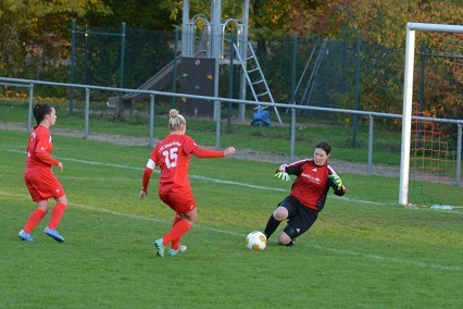 Zwei Spielerinnen in roten Trikots dribbeln auf das Tor zu, w&auml;hrend die Torh&uuml;terin in schwarz den Ball abwehrt.