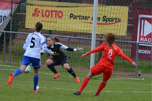 Zwei Fu&szlig;ballspielerinnen in rot und blau, w&auml;hrend ein Schuss auf das Tor abgefeuert wird, Torh&uuml;terin bereitet sich auf den Fang vor.