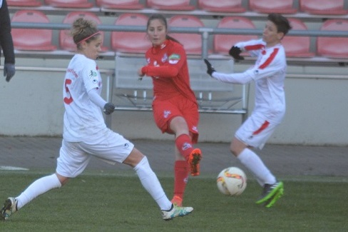 Spielszene einer Fu&szlig;ballerinnen: Eine Spielerin in Rot schie&szlig;t den Ball, w&auml;hrend zwei Spielerinnen in Wei&szlig; verteidigen.