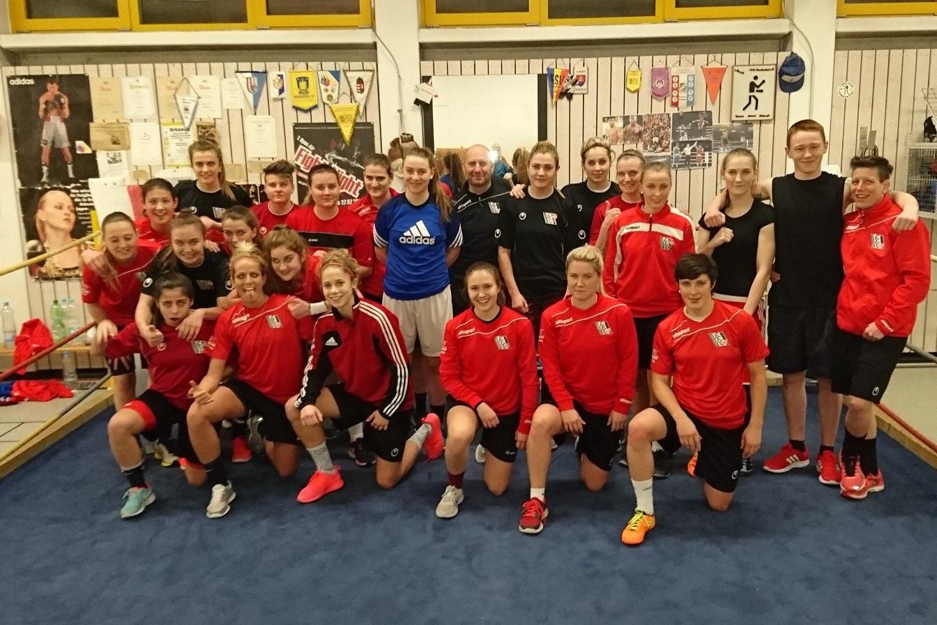 Gruppe von Sportlern in roten Trikots posiert in einem Trainingsraum, umgeben von Boxausr&uuml;stung und Plakaten.