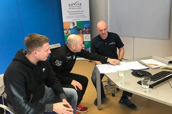 Drei M&auml;nner diskutieren &uuml;ber Dokumente an einem Tisch in einem modernen Besprechungsraum mit computergest&uuml;tzter Ausstattung.