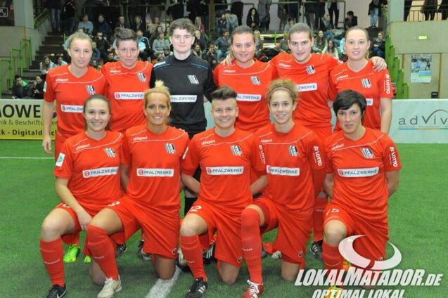 Gruppenfoto eines Frauenfu&szlig;ballteams in roten Trikots, stehend und kniend auf einem gr&uuml;nen Spielfeld.