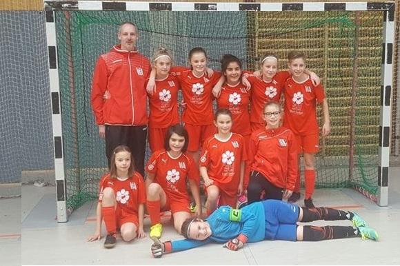 M&auml;dchen-Fu&szlig;ballmannschaft in roten Trikots posiert vor einem Tor, mit Trainer und Torh&uuml;ter in blauer Ausr&uuml;stung.