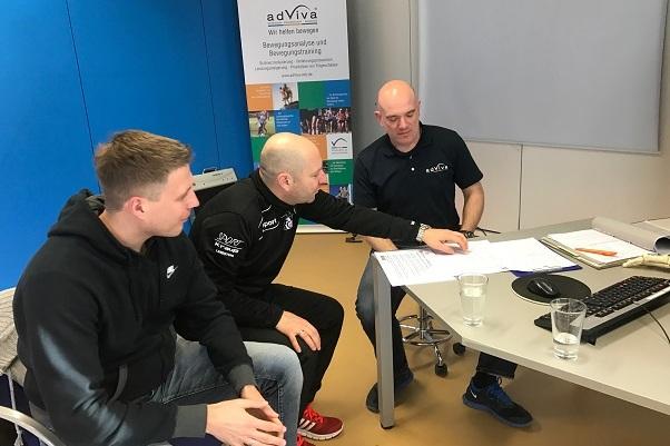 Drei M&auml;nner diskutieren &uuml;ber Dokumente an einem Tisch in einem modernen Besprechungsraum mit computergest&uuml;tzter Ausstattung.