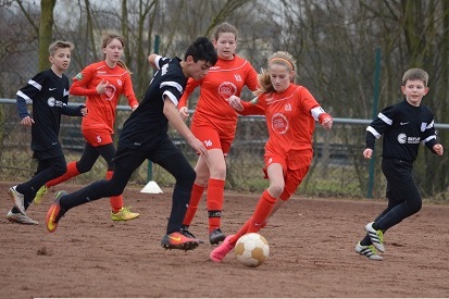 Jugendliche Fu&szlig;ballspieler in roten und schwarzen Trikots k&auml;mpfen um den Ball auf einem sandigen Spielfeld.