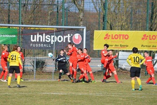 Torwart in schwarz h&auml;lt den Ball im Get&uuml;mmel von Spielerinnen in roten Trikots vor dem Tor. Gelbe Trikots im Hintergrund.