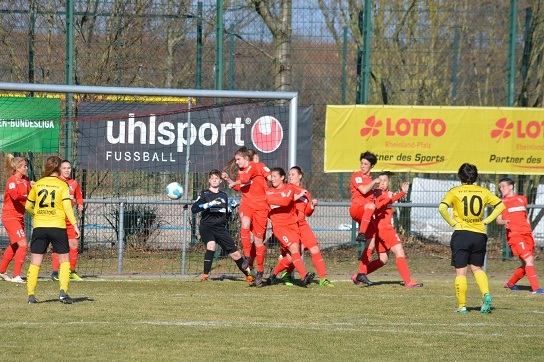 Torwart in schwarz h&auml;lt den Ball im Get&uuml;mmel von Spielerinnen in roten Trikots vor dem Tor. Gelbe Trikots im Hintergrund.