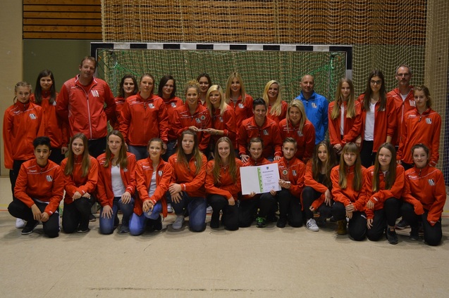 Gruppe von 24 Personen in roten Trainingsjacken, versammelt vor einem Handballtor, h&auml;lt ein Zertifikat in der Mitte.