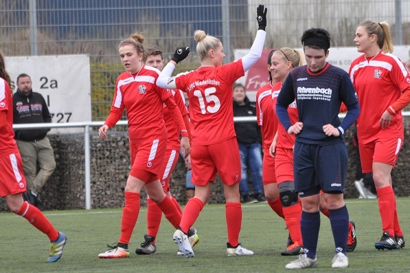 Frauenfu&szlig;ballmannschaft in roten Trikots jubelt auf dem Platz, w&auml;hrend eine Spielerin in blauer Kleidung zuschaut.