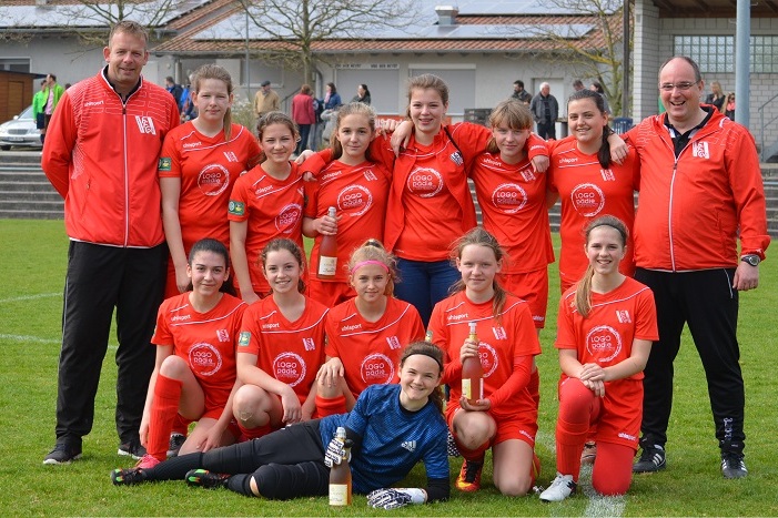 M&auml;dchenfu&szlig;ballmannschaft in roten Trikots posiert auf dem Rasen, Trainer hinter ihnen, Gewichte und Pokale in den H&auml;nden.