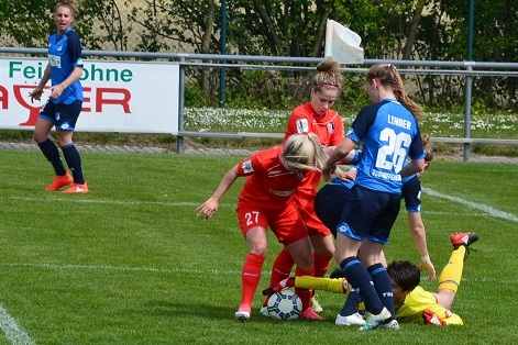 Zwei Fu&szlig;ballerinnen in roten Trikots k&auml;mpfen um den Ball auf einem gr&uuml;nen Fu&szlig;ballfeld, eine Torh&uuml;terin liegt am Boden.