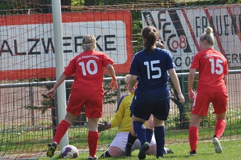 Spielszene im Fu&szlig;ball mit zwei Spielerinnen in roten Trikots und einer in blau, die auf den Ball vor dem Torwart zustreben.
