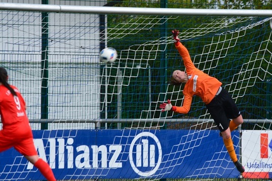 Torwart in orange springt nach oben, um einen Fu&szlig;ball entschlossen zu parieren, w&auml;hrend eine Spielerin in Rot zuschaut.