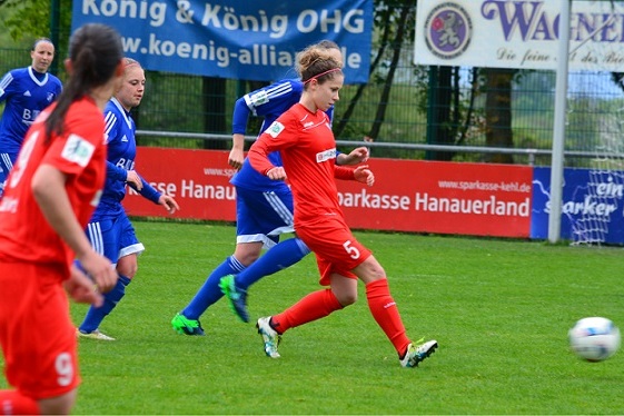 Frauenfu&szlig;ballspiel: Spielerin in rotem Trikot dribbelt mit Ball, w&auml;hrend zwei Spielerinnen in blauen Trikots verfolgen.