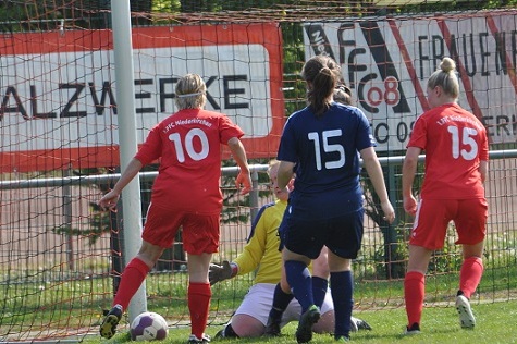 Spielszene im Fu&szlig;ball mit zwei Spielerinnen in roten Trikots und einer in blau, die auf den Ball vor dem Torwart zustreben.