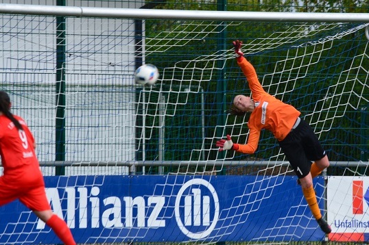 Torwart in orange springt nach oben, um einen Fu&szlig;ball entschlossen zu parieren, w&auml;hrend eine Spielerin in Rot zuschaut.
