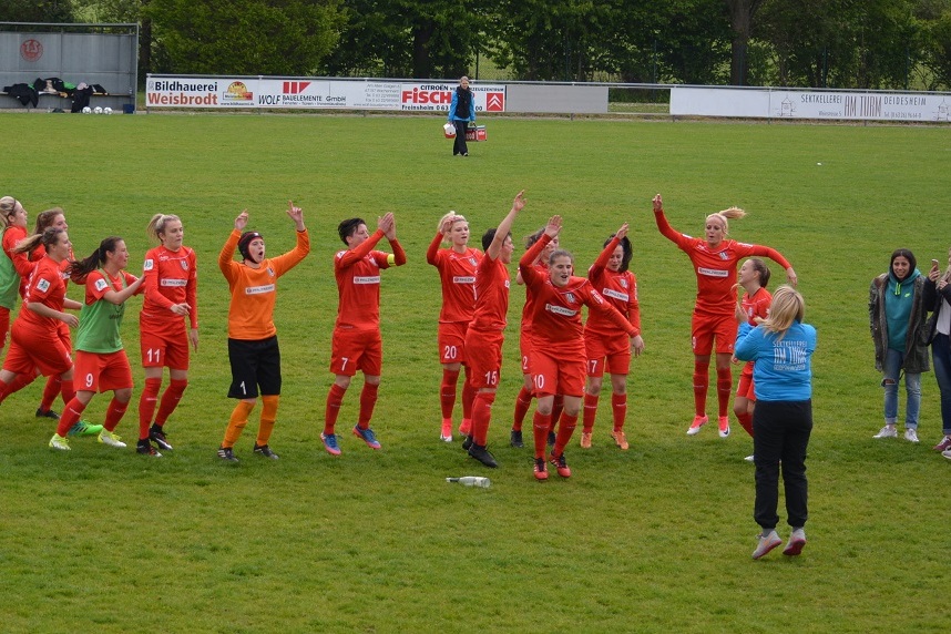 Frauenfu&szlig;ballmannschaft in roten Trikots jubelt auf dem Spielfeld, w&auml;hrend Zuschauer am Rand stehen.