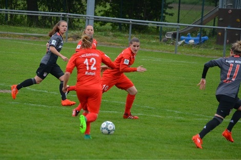 Vier Fu&szlig;ballspielerinnen in roten und grauen Trikots spielen auf einem Rasenfeld, w&auml;hrend der Ball in Richtung des roten Teams rollt.