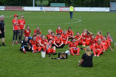 Gruppe von weiblichen Fu&szlig;ballspielerinnen in roten Trikots sitzt auf dem Rasen und freut sich gemeinsam.